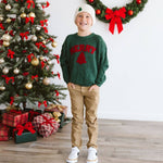 Child wearing a green sweater with red text standing in front of a decorated Christmas tree.