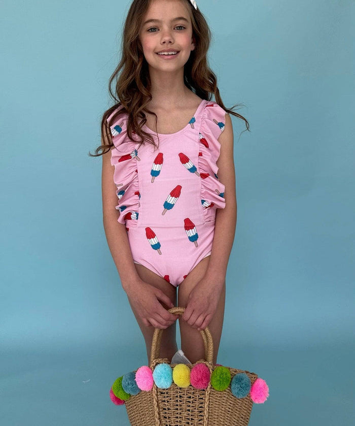 Young girl wearing a pink swimsuit with a popcicle design, holding a colorful pom-pom bag against a blue background.
