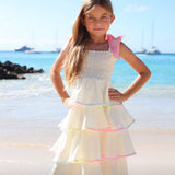 Young girl in a white dress with colorful trim standing on a beach.