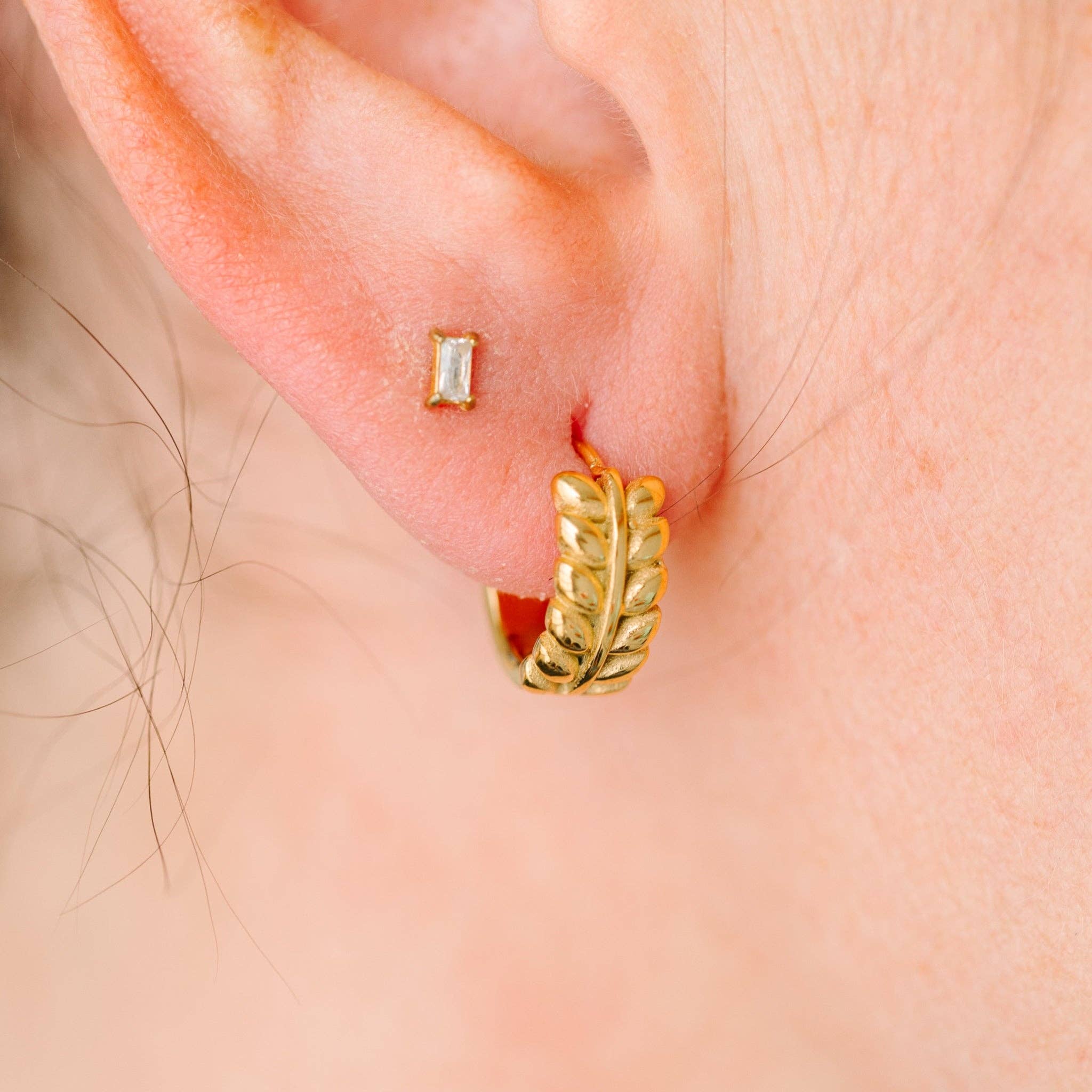 Close-up of an ear with two gold earrings: a rectangular one and a leaf-shaped one.