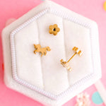 Gold unicorn-shaped earrings on a white hexagonal stand against a pink background