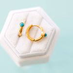 Two gold earrings with turquoise stones on a white hexagonal jewelry box against a light blue background