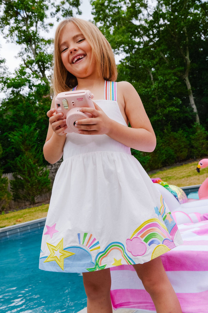 Young girl in a white dress with colorful designs by a pool holding a pink phone.