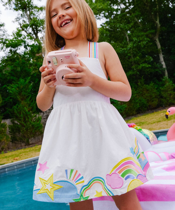 Young girl in a white dress with colorful designs by a pool holding a pink phone.