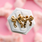 Gold bow-shaped earrings on a hexagonal white stand with a pink floral background