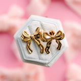Gold bow-shaped earrings on a hexagonal white stand with a pink floral background
