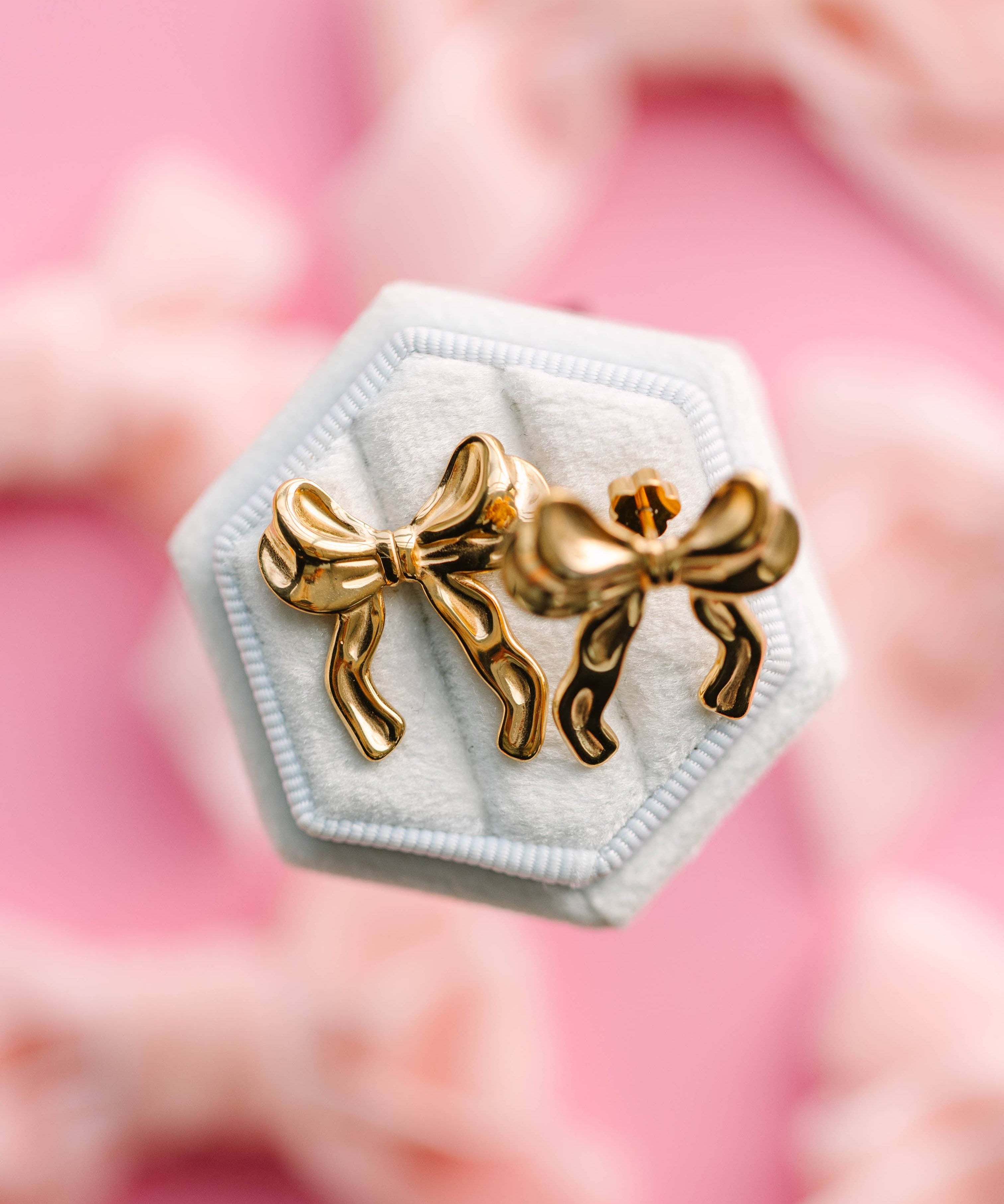 Gold bow-shaped earrings on a hexagonal white stand with a pink floral background