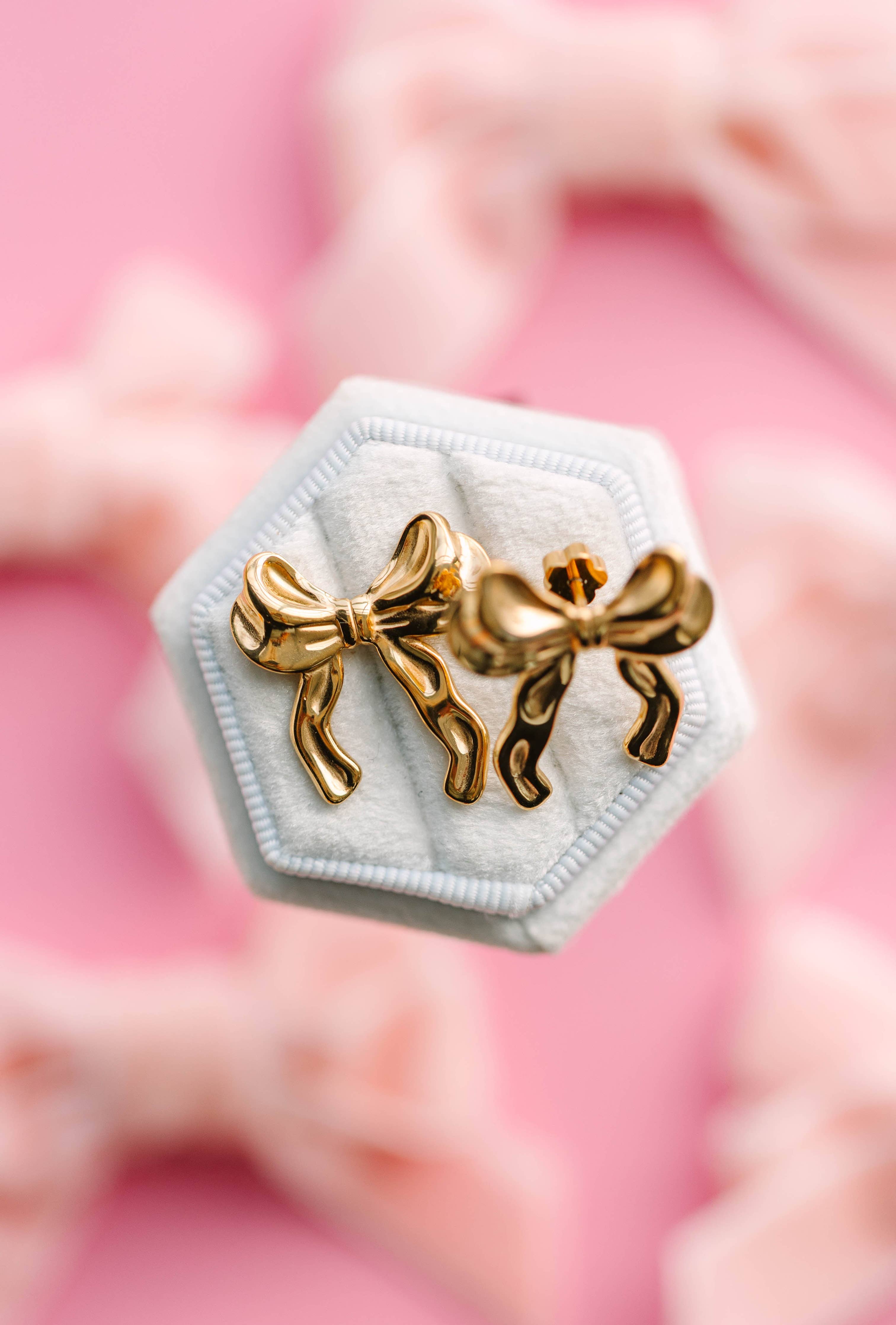 Gold bow-shaped earrings on a hexagonal white stand with a pink floral background