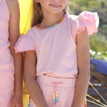 Two young girls in matching pink outfits with rainbow accents standing on a beach.