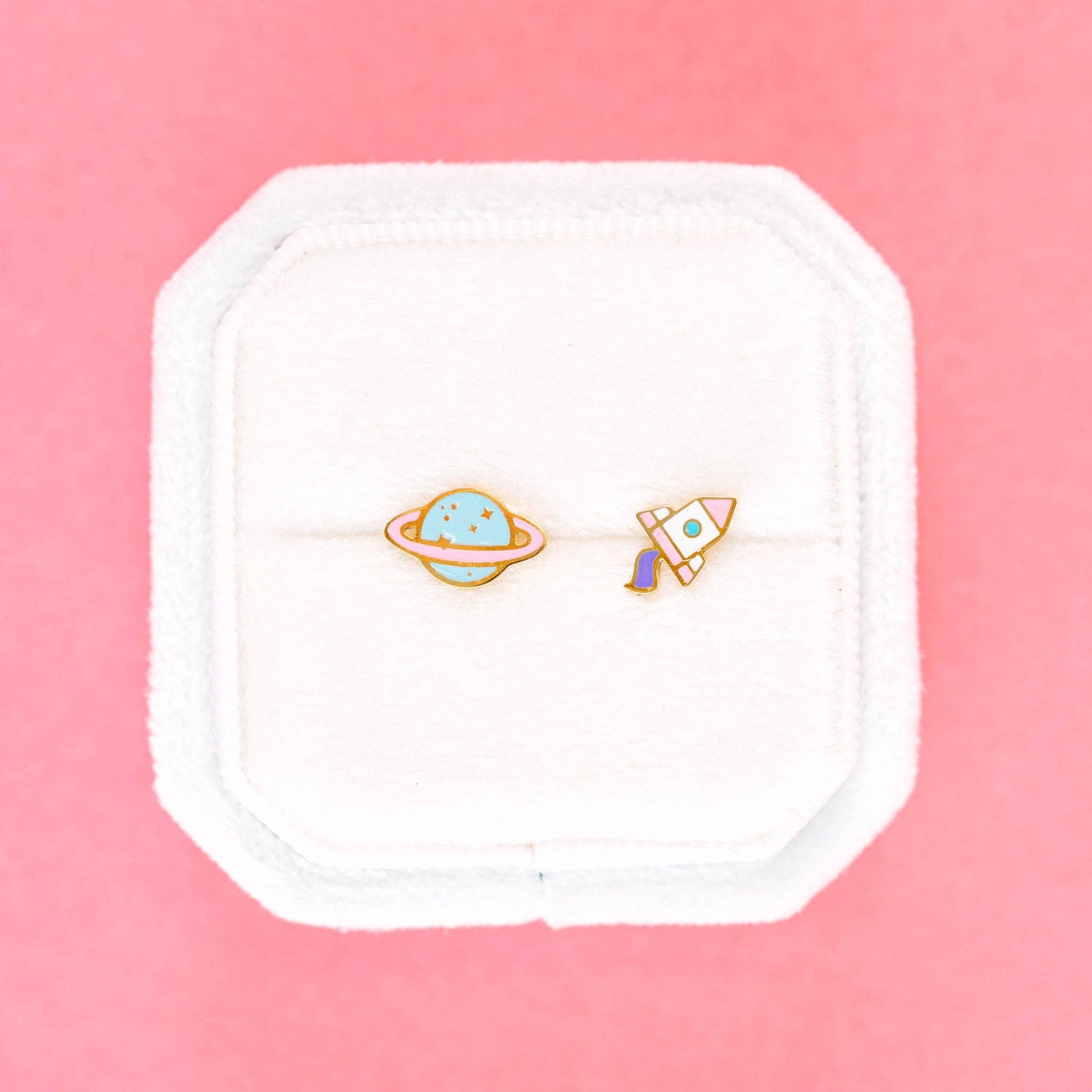 space earrings, one planet and one rocket ship on a cushion with prink background