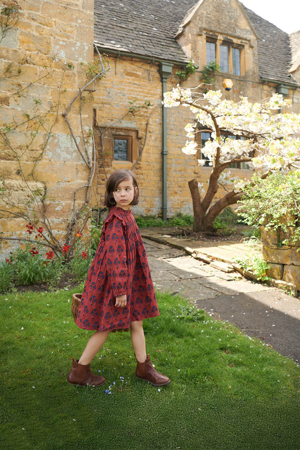 Young girl in a red dress standing in front of a stone building with a tree in the background.