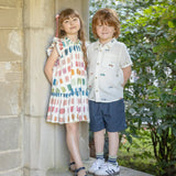 Two children standing outdoors, one in a colorful dress and the other in a light shirt and navy shorts.