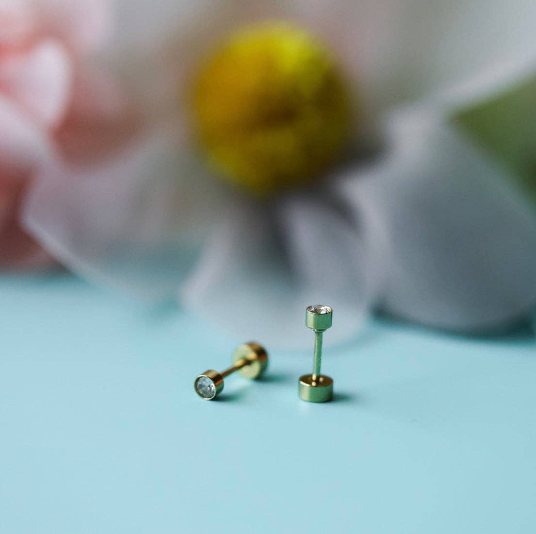 Gold stud earrings on a light blue surface with a blurred floral background