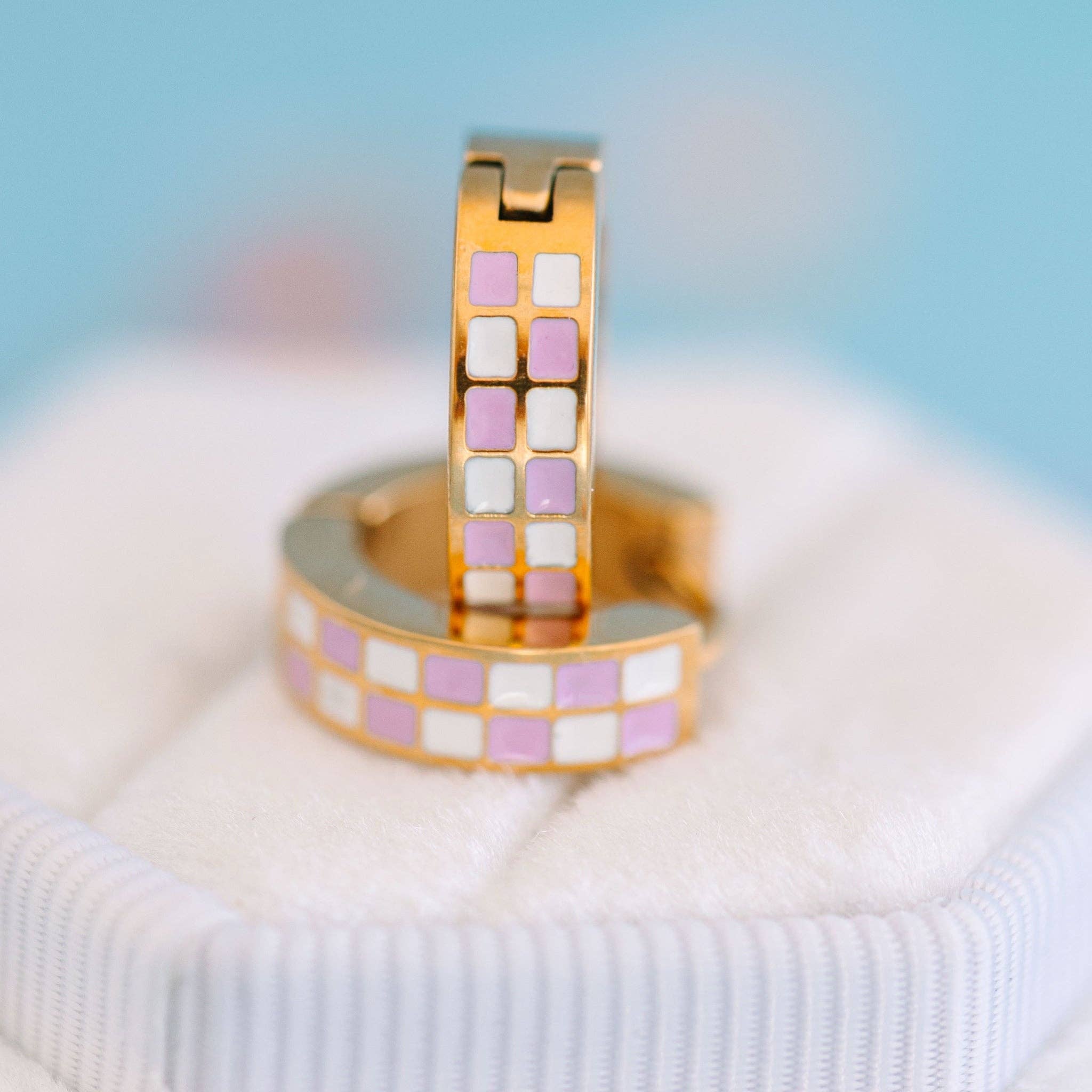 Gold earrings with lilac and white checkered pattern on a soft fabric background
