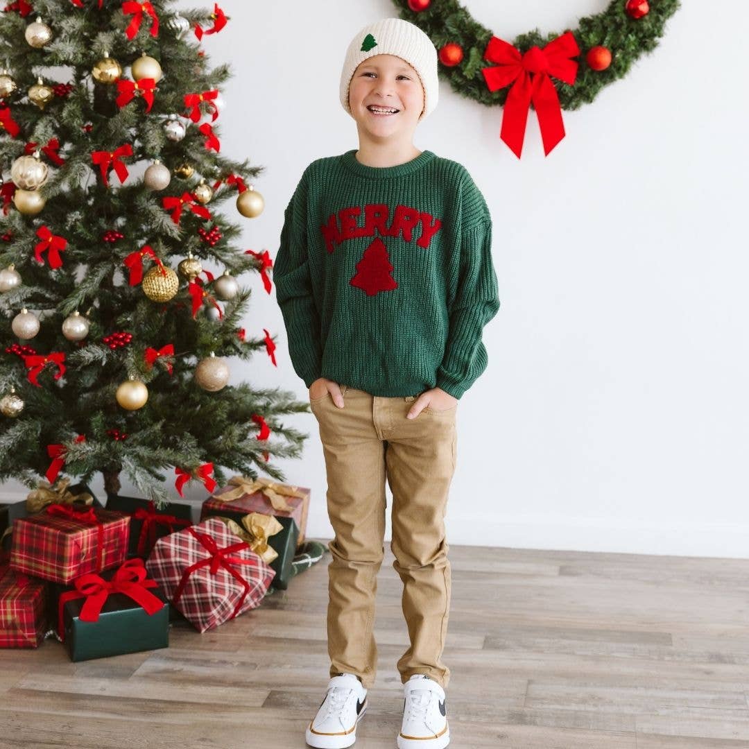 Child wearing a green sweater with red text standing in front of a decorated Christmas tree.