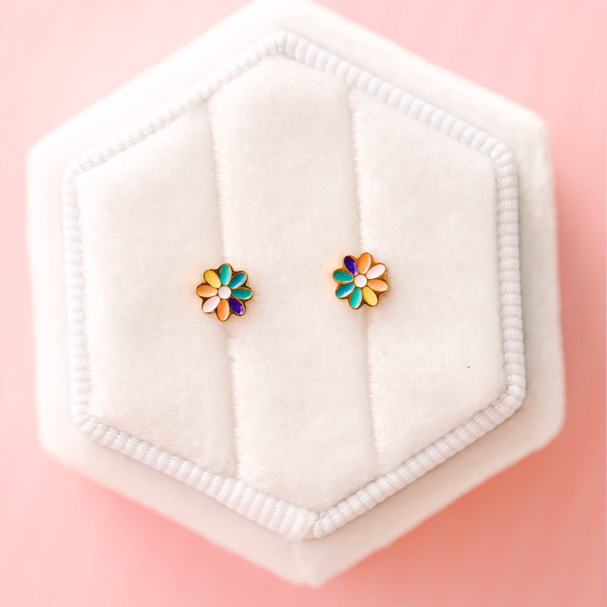 Colorful flower-shaped earrings on a white hexagonal stand against a pink background