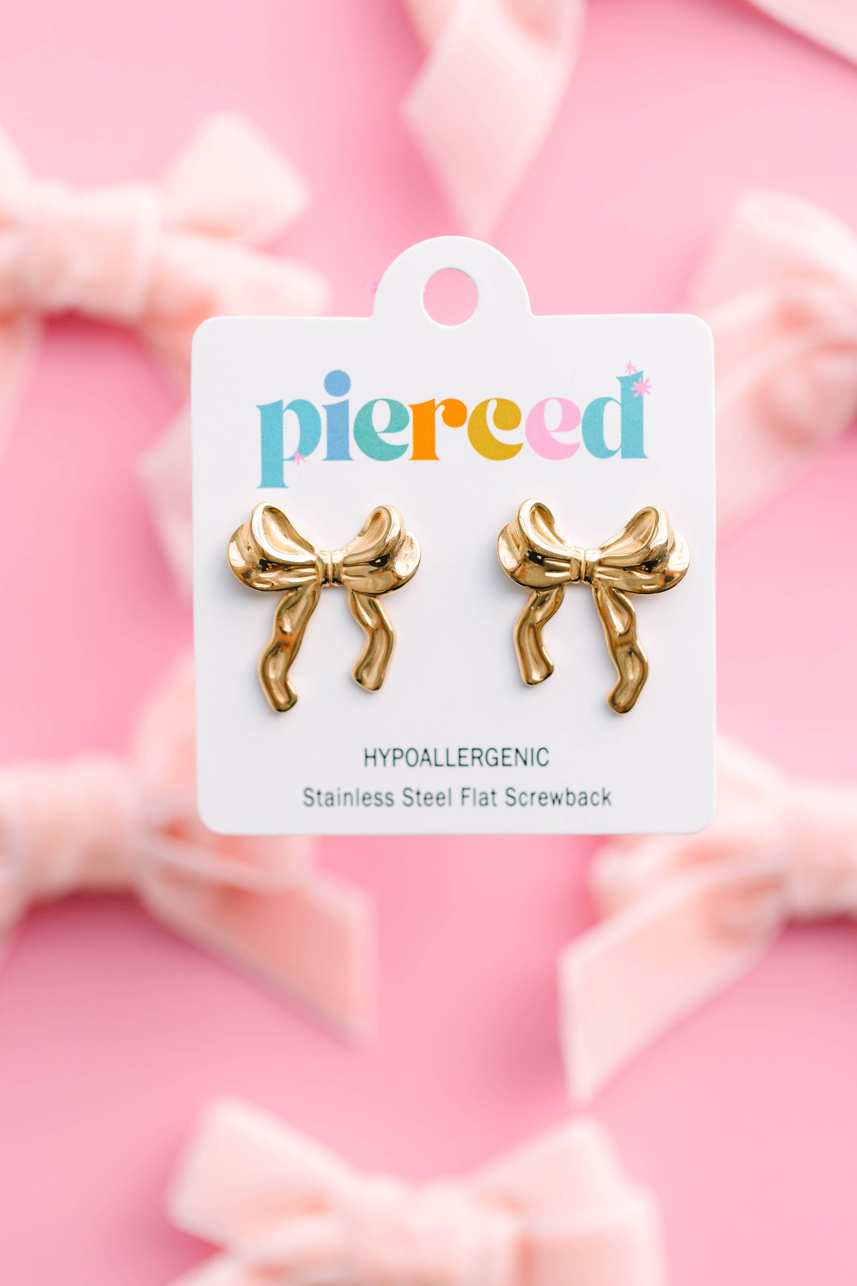Gold bow-shaped earrings on a 'pierced' branded card against a pink background