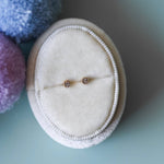 Pair of gold earrings on a white round cushion with colorful pom-poms in the background.